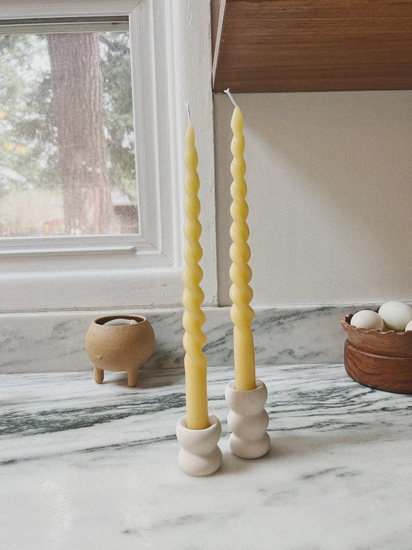 Two yellow spiral candles in white holders on a marble surface with a window in the background.