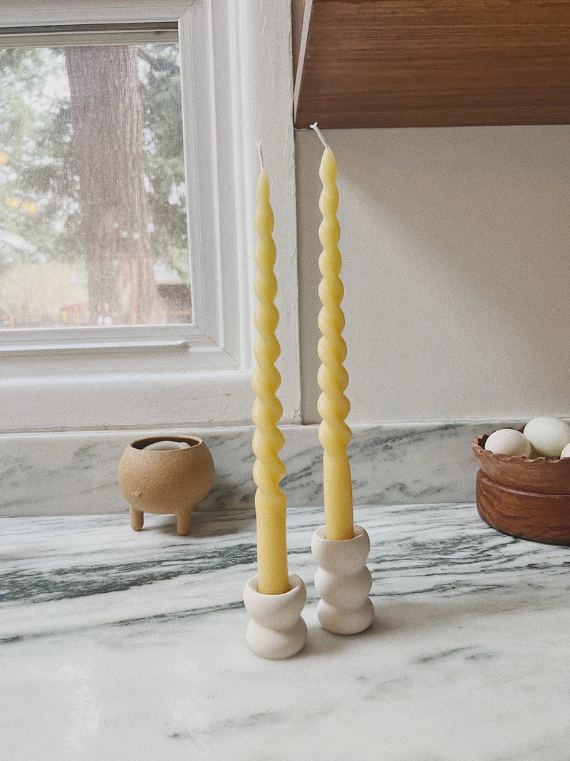 Two yellow spiral candles in white holders on a marble surface with a window in the background.