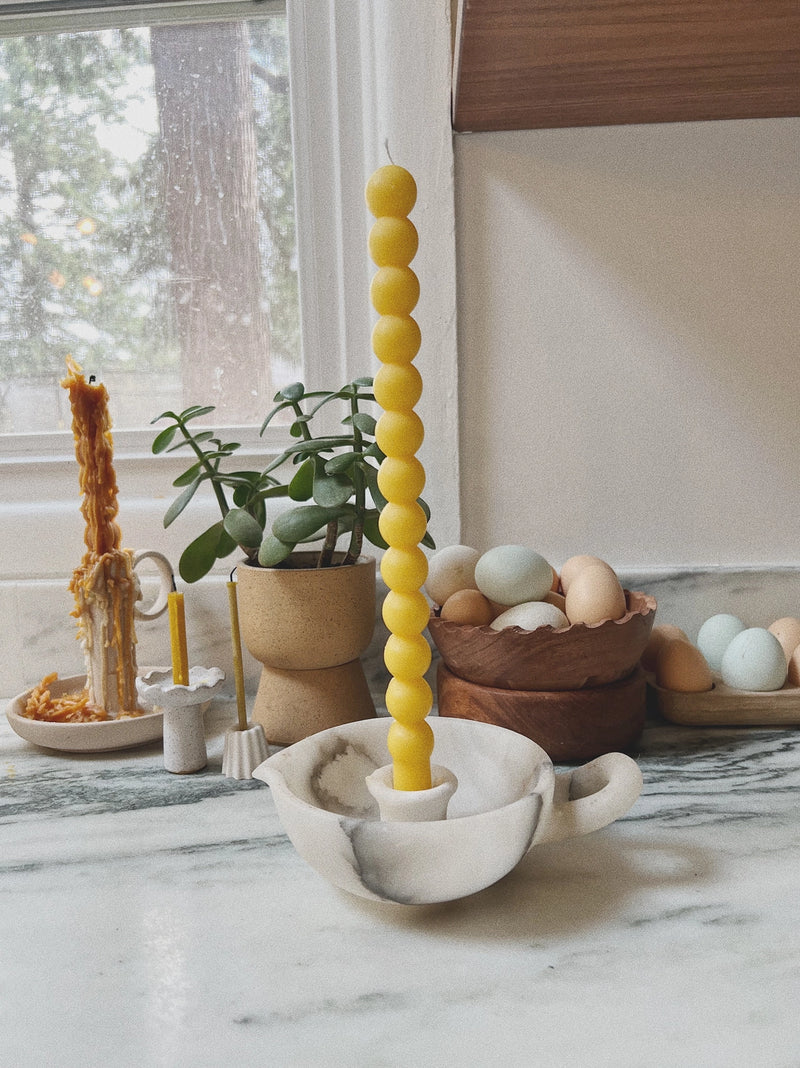 Yellow candle in a marble holder on a marble surface with plants and eggs in the background.