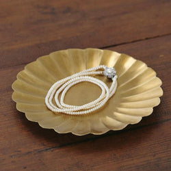 Brass jewelry dish with a pearl necklace on a wooden surface.

Fog Linen round petal brass plate tray dish serving catch all home decor kitchen Boston gift shop store boutique