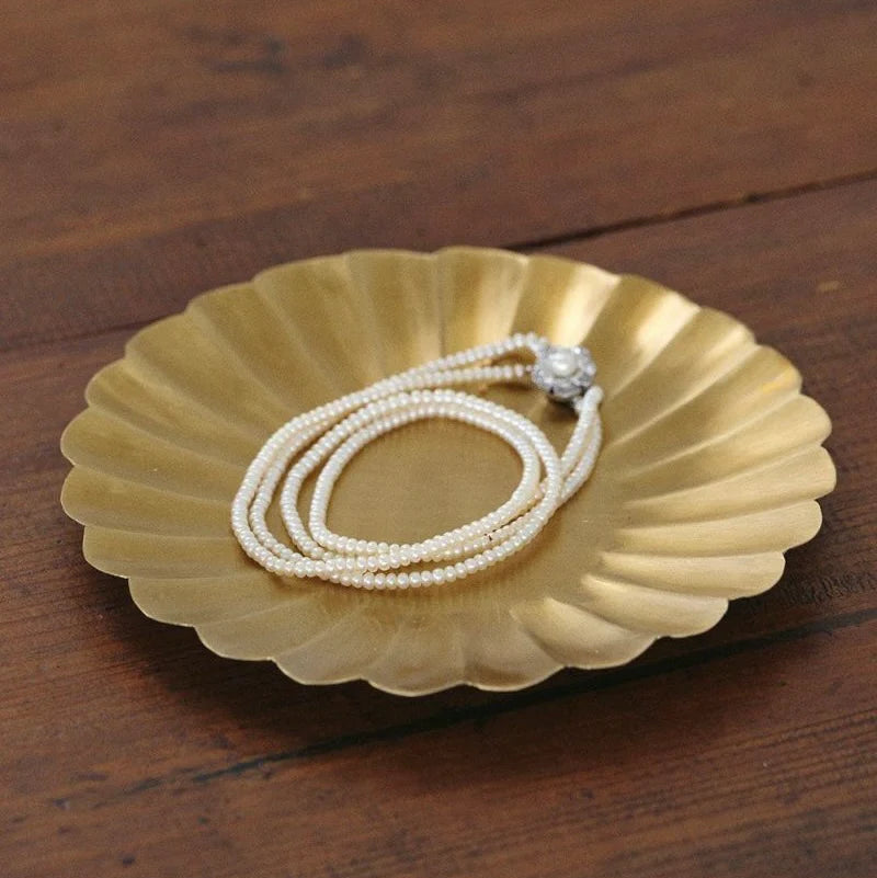 Brass jewelry dish with a pearl necklace on a wooden surface.

Fog Linen round petal brass plate tray dish serving catch all home decor kitchen Boston gift shop store boutique