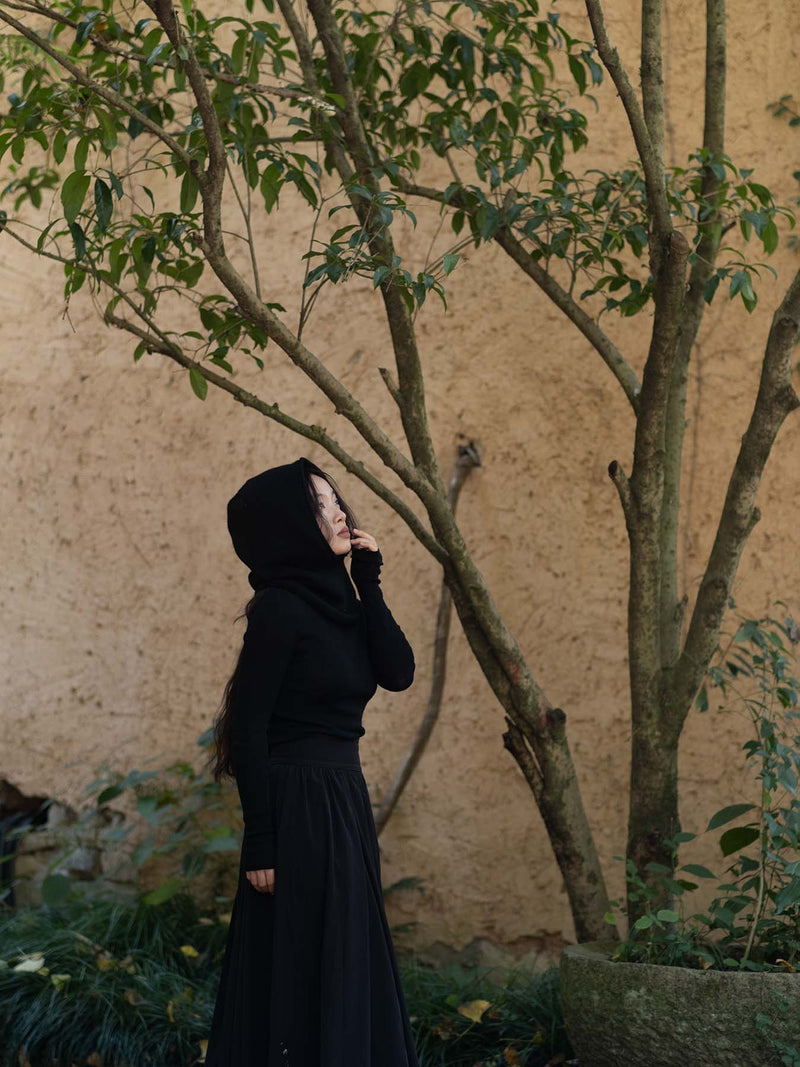 Person wearing a black hood and black outfit standing in front of a textured wall with trees.