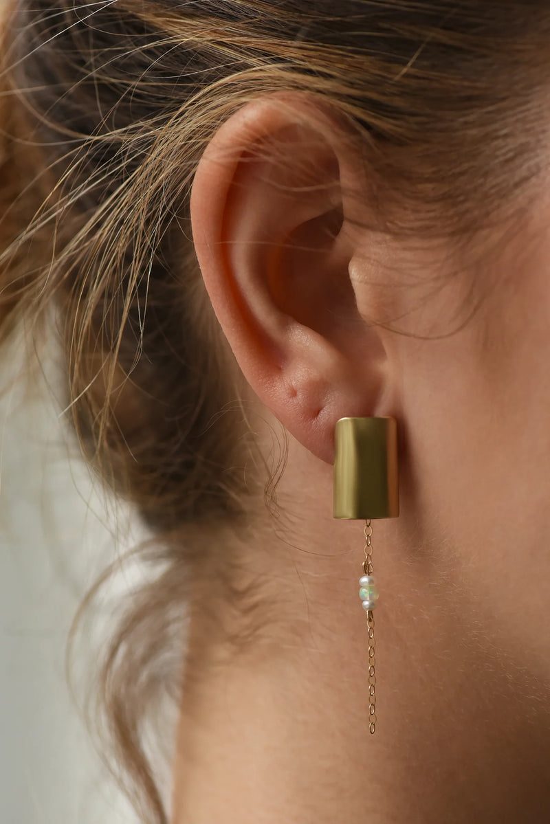 Close-up of a brass earring with a chain and opal and pearl beads worn by a person with blonde hair.