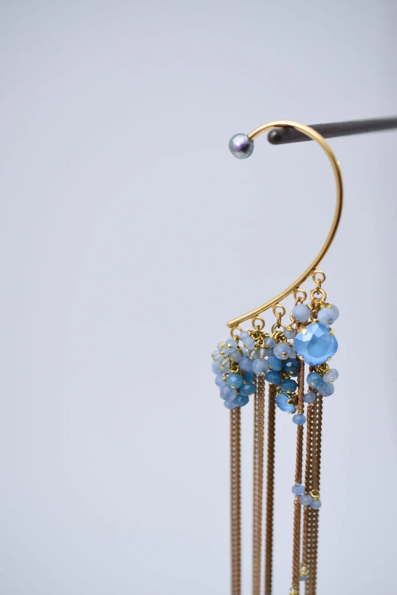 Close-up of a person wearing a ear cuff with brass chain fringe and blue stones on a white background