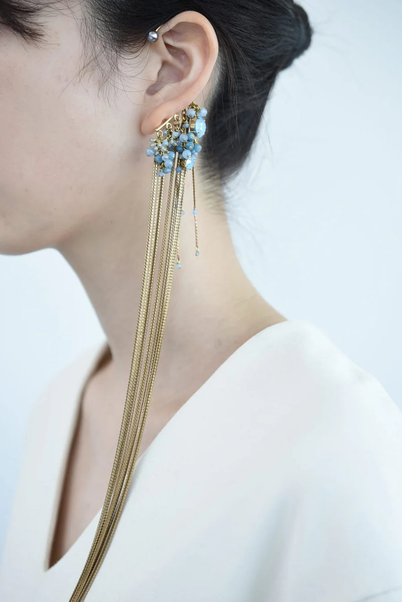 Close-up of a person wearing a ear cuff with brass chain fringe and blue stones on a white background