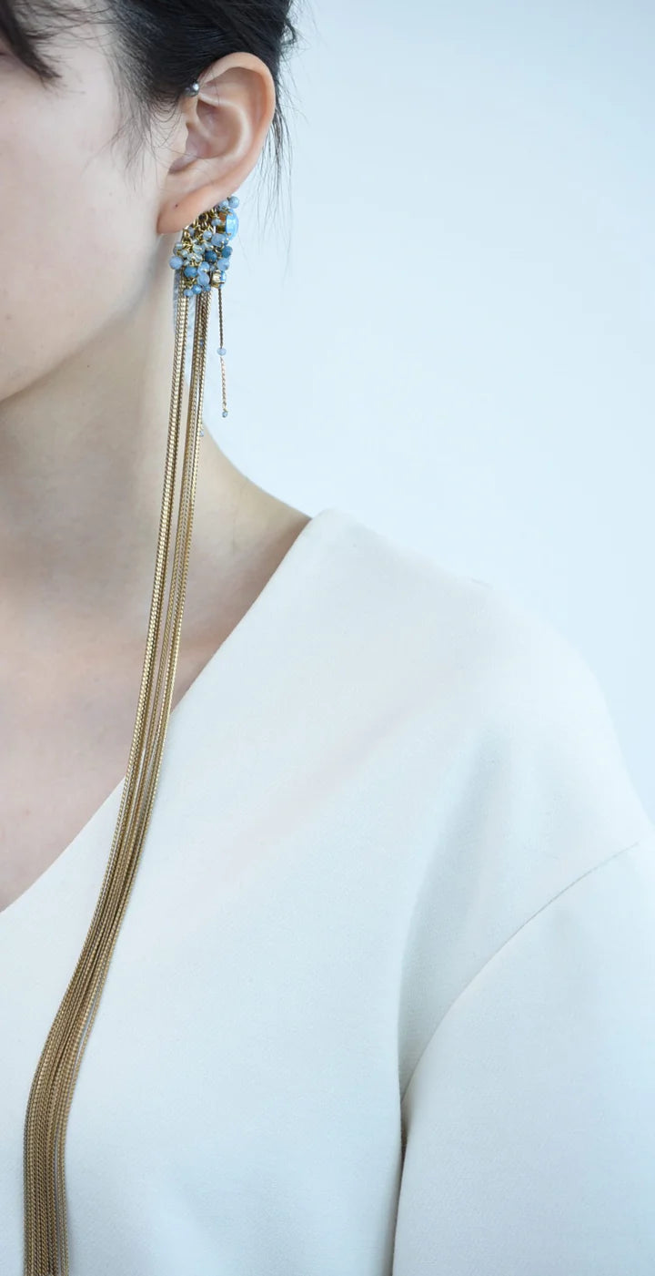 Close-up of a person wearing a ear cuff with brass chain fringe and blue stones on a white background