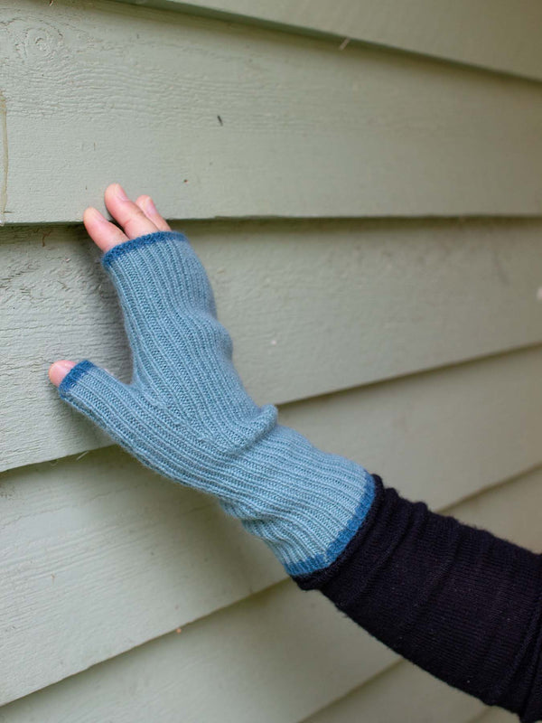 Blue knitted fingerless glove on a hand against a beige wall.