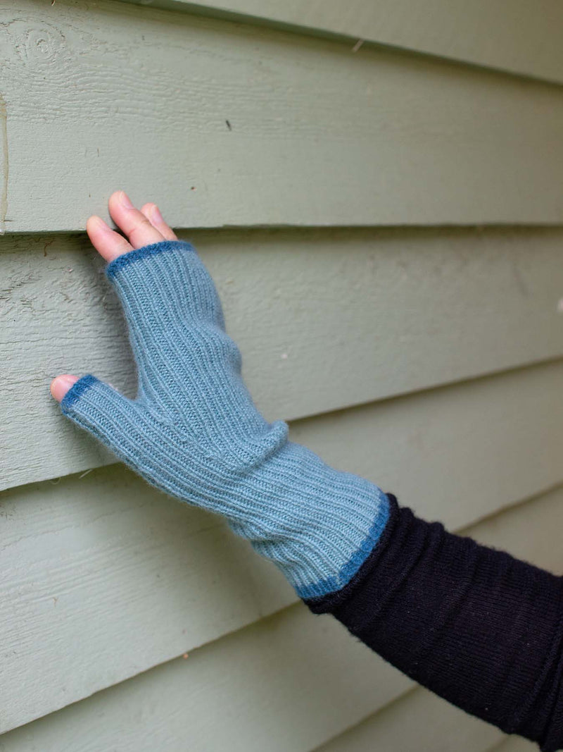 Blue knitted fingerless glove on a hand against a beige wall.