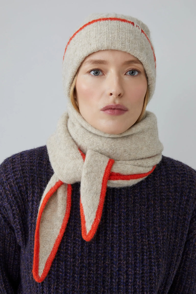 Person wearing a beige knit hat and scarf with orange accents on a plain background