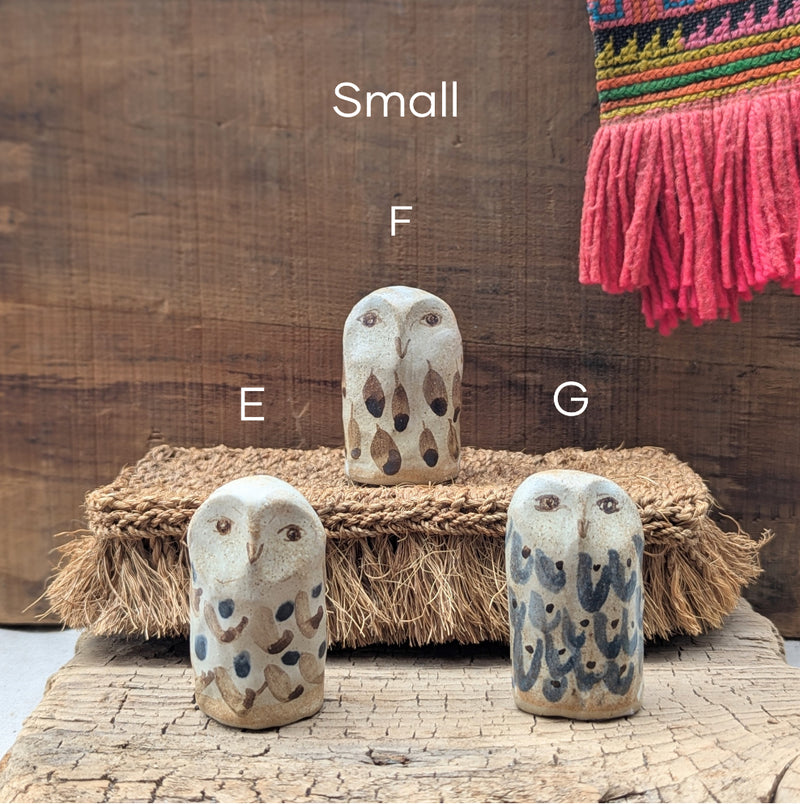 Three ceramic owl figurines size small on a wooden surface with a textured wall and colorful fabric in the background.