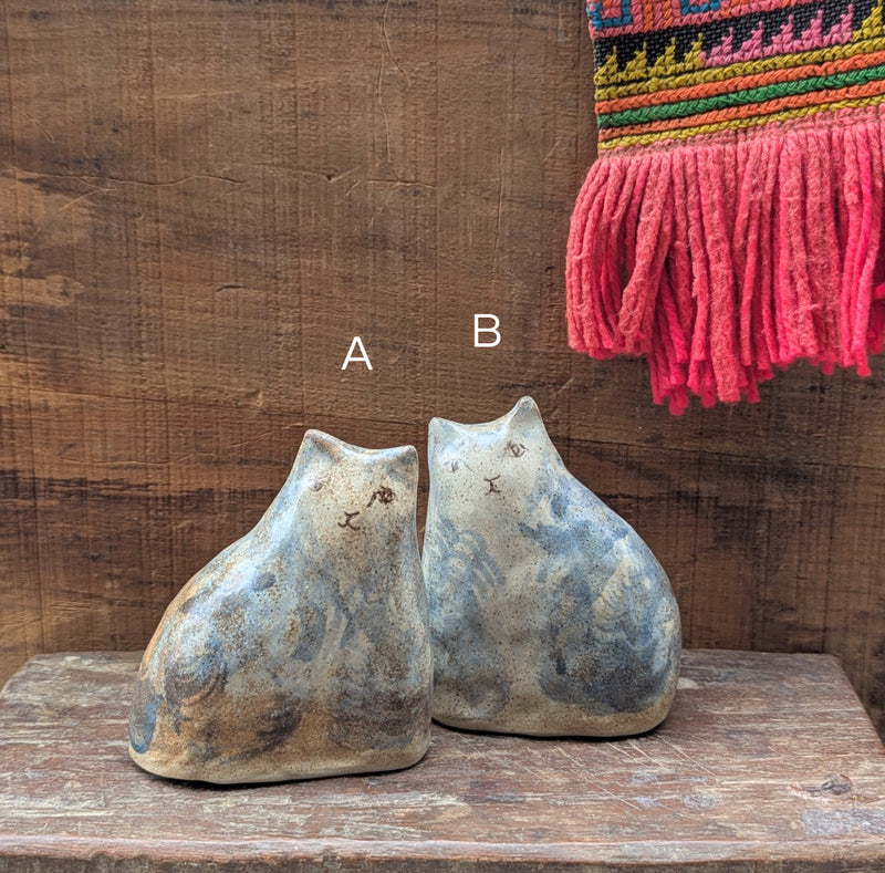 Two ceramic cat figurines on a wooden surface with a colorful woven textile in the background.