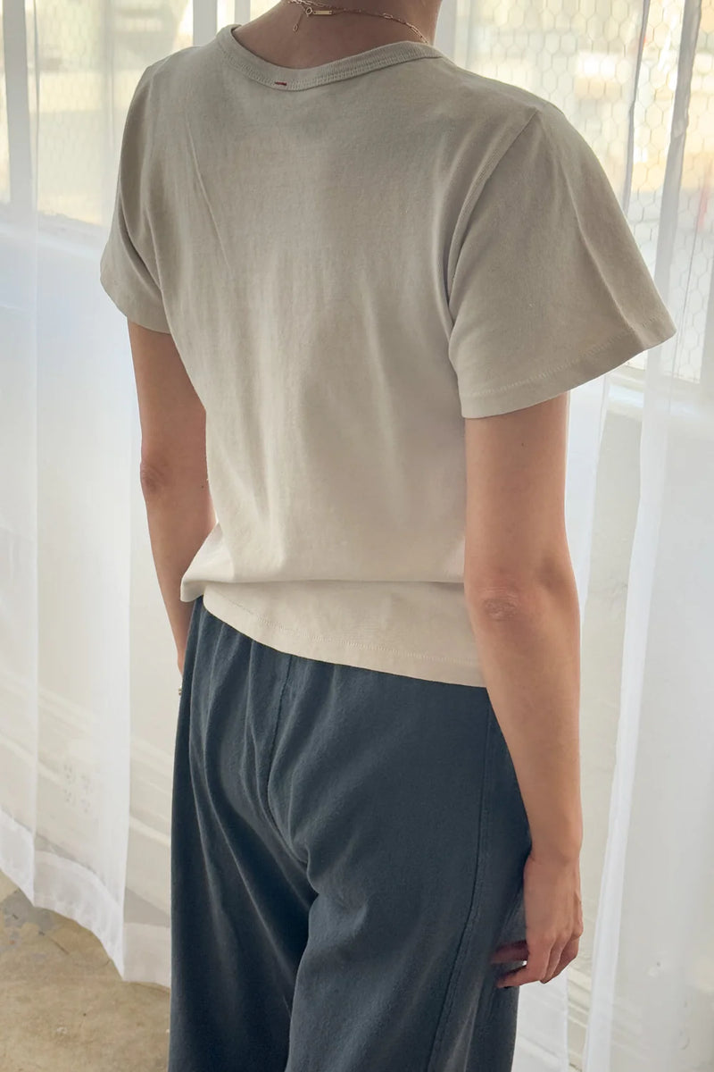 Person wearing a light beige t-shirt and dark pants from Le Bon Shoppe standing in front of sheer curtains.