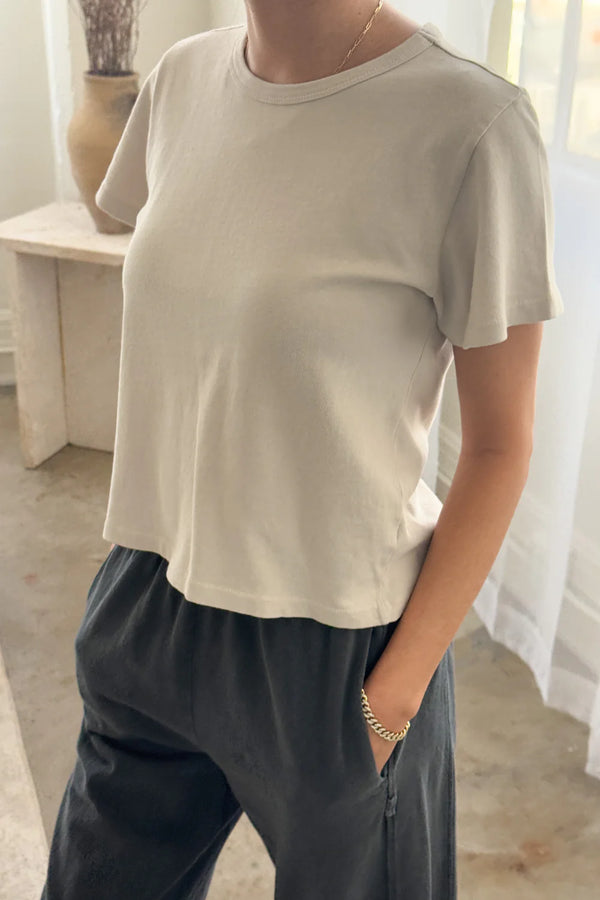 Person wearing a light beige t-shirt and dark pants from Le Bon Shoppe standing in front of sheer curtains.