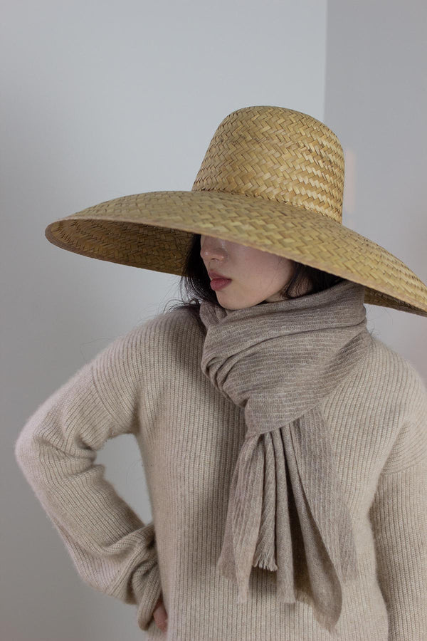 Person wearing a wide-brimmed straw hat and beige sweater with a scarf against a plain background