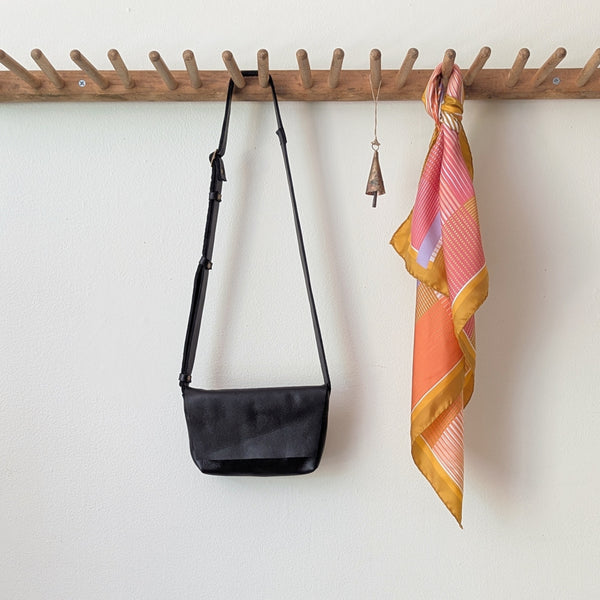 Black bag and colorful scarf hanging on a wooden rack against a white wall. Stitch and Tickle handmade leather mini messenger bag crossbody made in Boston made in usa leatherwork leathercraft SoWa shop boutique gift store