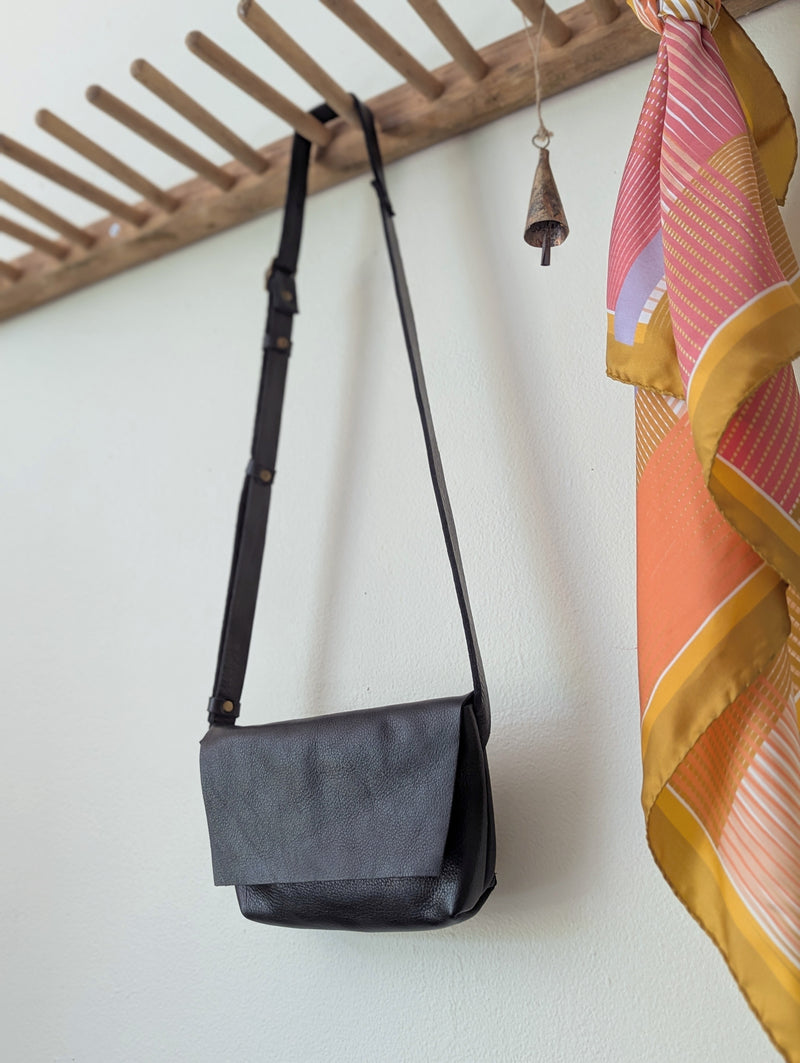 Black shoulder bag hanging on a wooden rack. Stitch and Tickle handmade leather mini messenger bag crossbody made in Boston made in usa leatherwork leathercraft SoWa shop boutique gift store