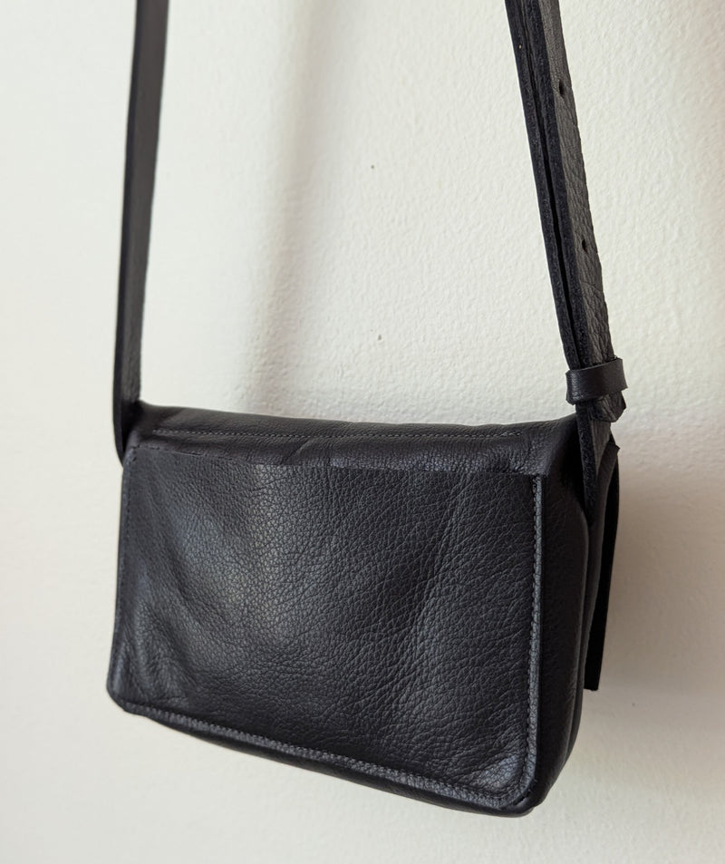Black leather crossbody bag on a light beige background. Stitch and Tickle handmade leather mini messenger bag crossbody made in Boston made in usa leatherwork leathercraft SoWa shop boutique gift store