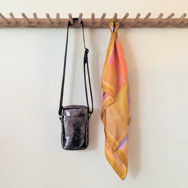 Small lack metallic crossbody bag and colorful scarf hanging on a wooden rack against a white wall. 
Stitch and Tickle unisex messenger travel bag adjustable strap shoulder crossbody leatherwork handmade sowa shop Boston gift shop store