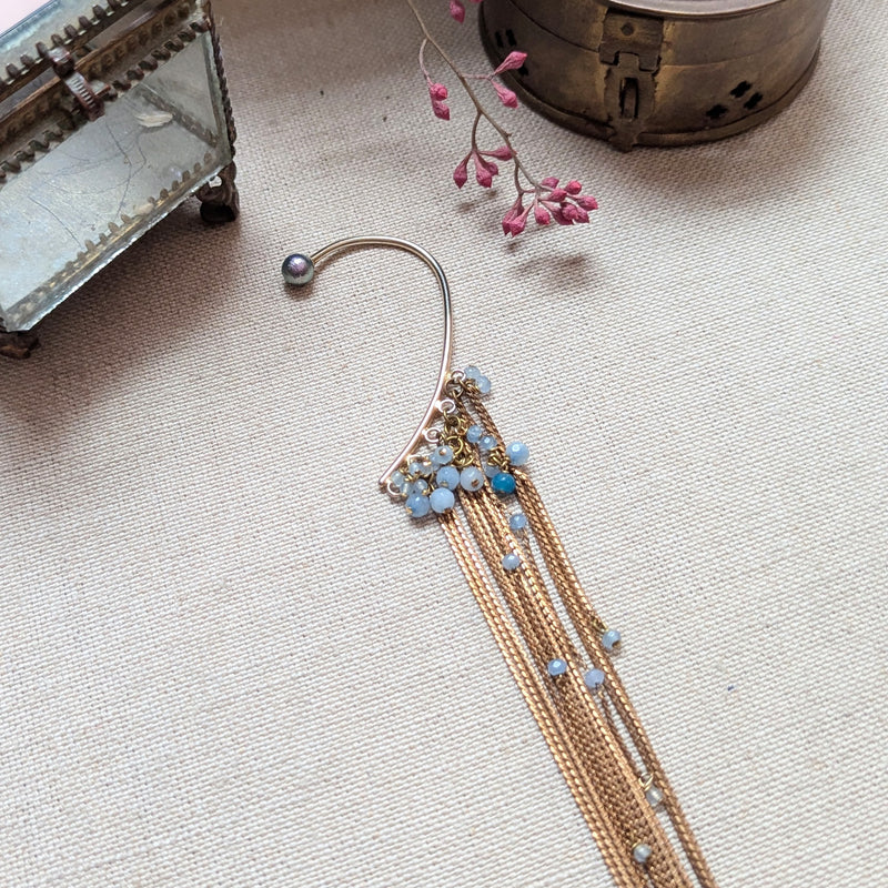 Brass ear cuff or ear hook with long  brass chain fringe and blue beads on a textured surface