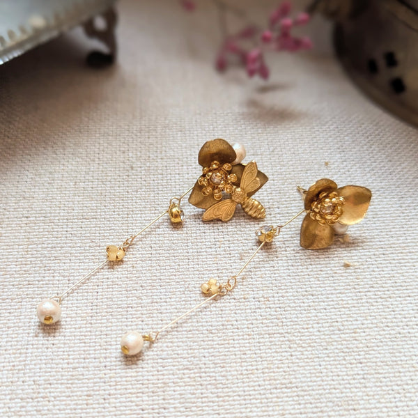 Brass flower and bee dangle earrings with pearl and crystal accents on a textured surface