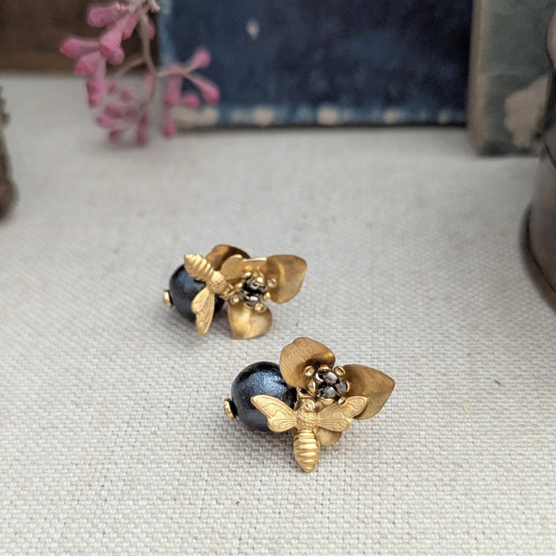 Brass bee and begonia blossom earrings on a texutred linen surface with a blurred background