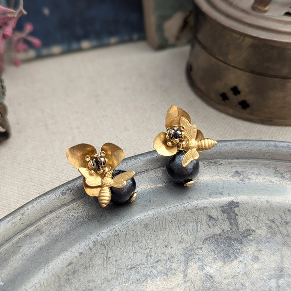 Brass bee and begonia blossom earrings on a metallic surface with a blurred background