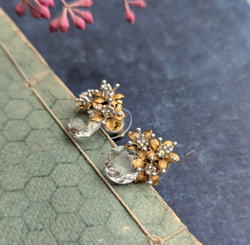 Pair of brass and crystal earrings on a textured surface with pink flowers.