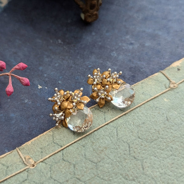 Pair of brass and crystal earrings on a textured surface with pink flowers.