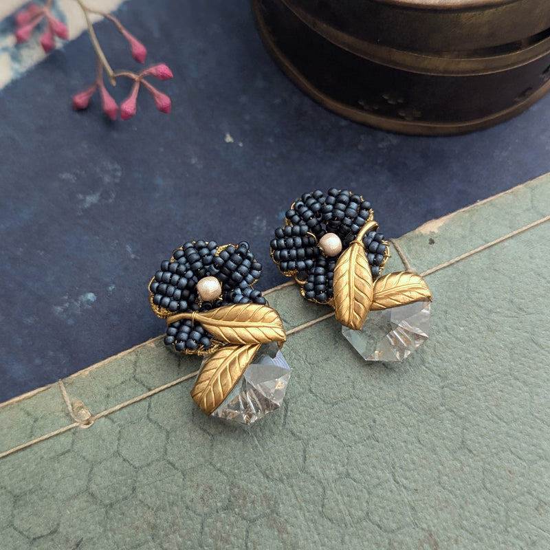 Pair of earrings with blue beads, gold leaves, and clear crystals on a textured surface.