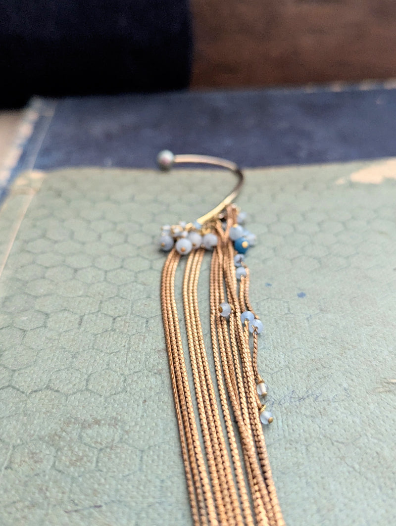Brass ear cuff or ear hook with long  brass chain fringe and blue beads on a textured surface