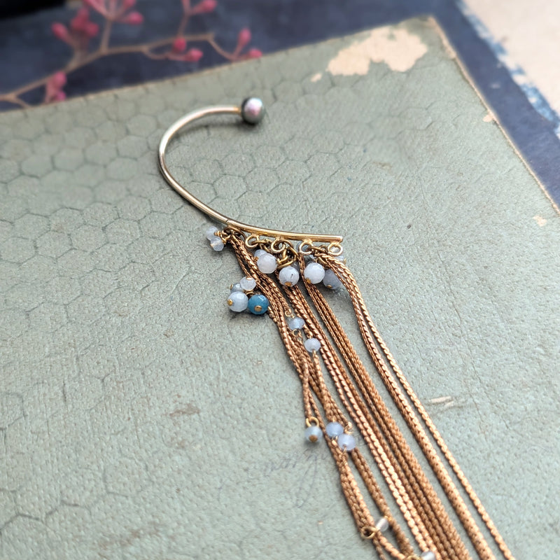 Brass ear cuff or ear hook with long  brass chain fringe and blue beads on a textured surface