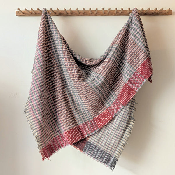 Plaid blanket with red and gray pattern draped over a wooden rack against a white wall.
