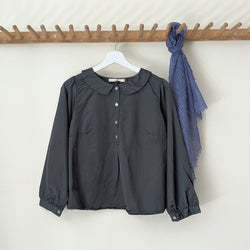 Dark green blouse with a collar  and buttons on a hanger next to a blue scarf on a wooden rack against a white wall.