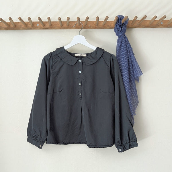 Dark green blouse with a collar  and buttons on a hanger next to a blue scarf on a wooden rack against a white wall.