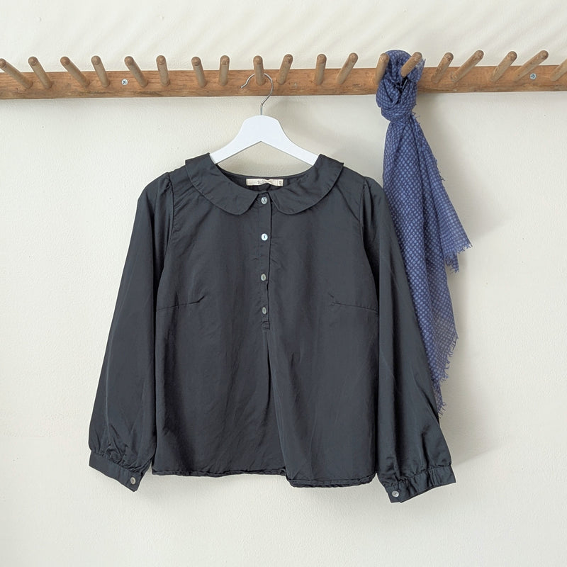 Dark green blouse with a collar  and buttons on a hanger next to a blue scarf on a wooden rack against a white wall.