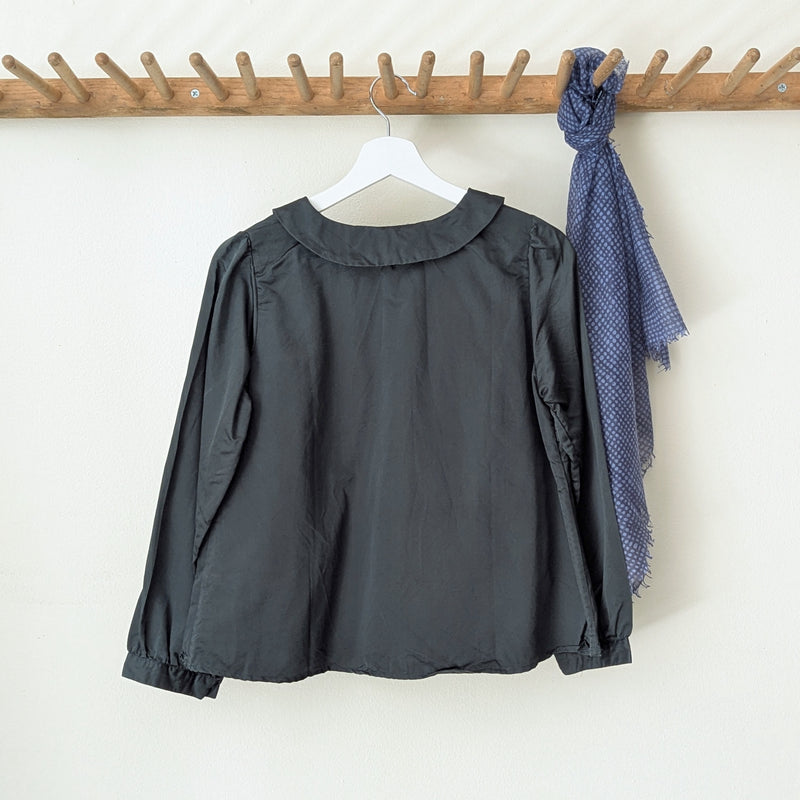 Dark green blouse with a collar  and buttons on a hanger next to a blue scarf on a wooden rack against a white wall.