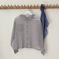 Sheer gray silk organza blouse with a blue patterned scarf on a wooden rack against a white wall.