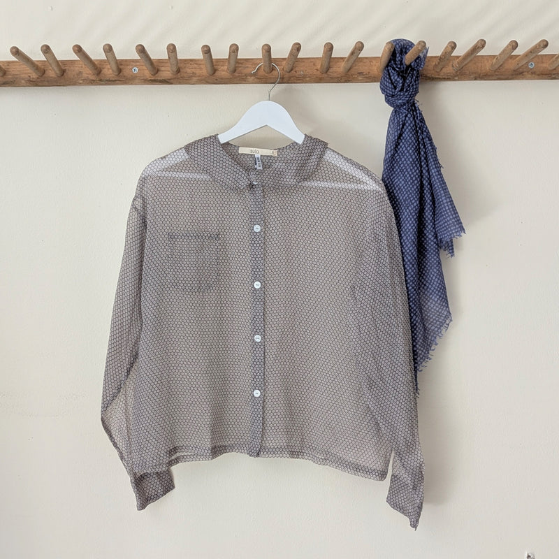 Sheer gray silk organza blouse with a blue patterned scarf on a wooden rack against a white wall.