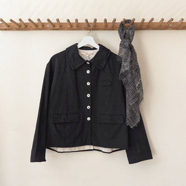 Black jacket with white buttons on a hanger against a beige wall.