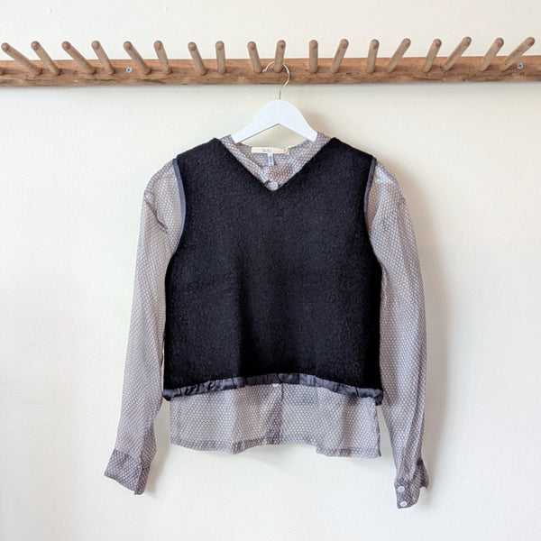 Black vest with gray layered shirt on a hanger against a white wall with a wooden rack above.