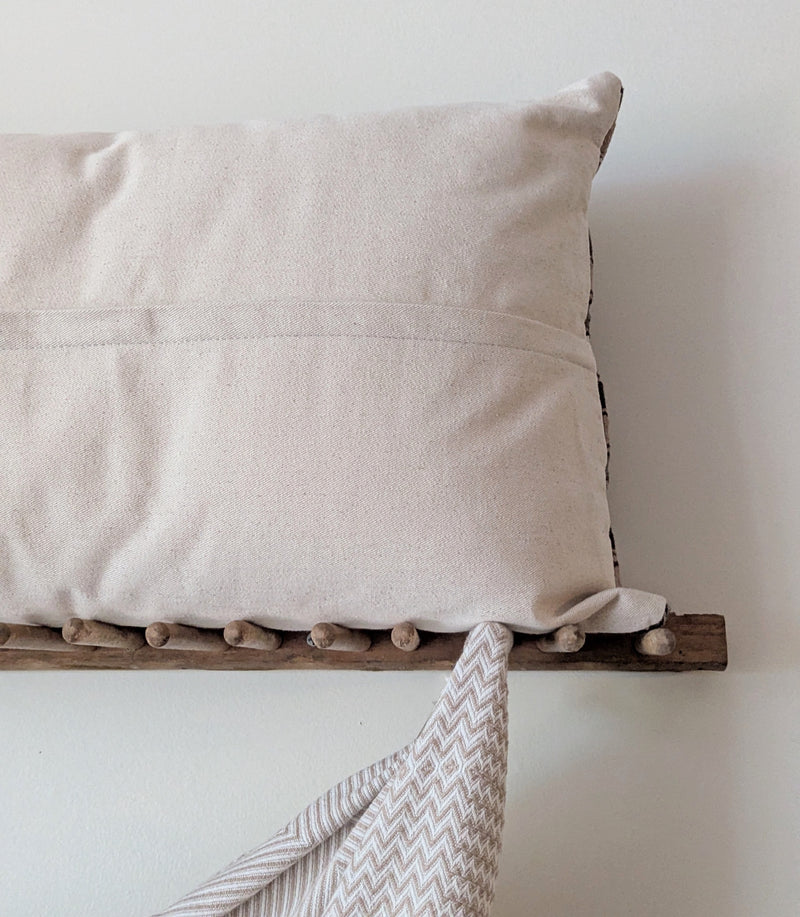 Plain beige back of a sriped pillow with a hidden zipper on a wooden rack with a draped blanket against a plain wall