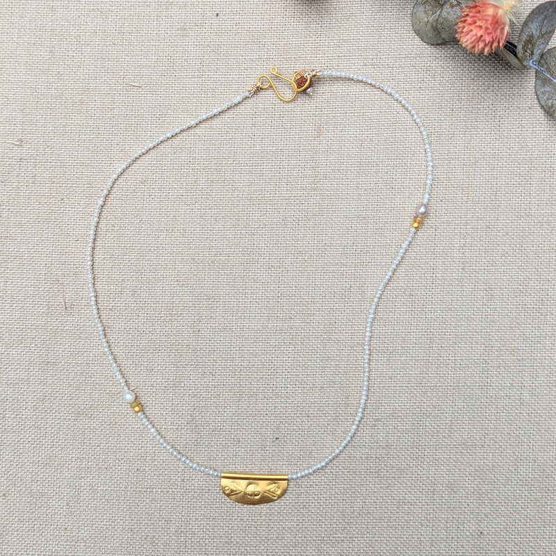Necklace with a half oval gold pendant with a textured design on a pearl beaded cord on a textured surface