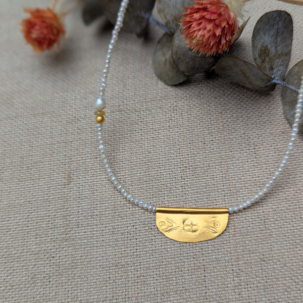 Necklace with a half oval gold pendant with a textured design on a pearl beaded cord on a textured surface with flowers and leaves.