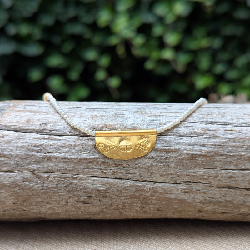Necklace with a half oval gold pendant with a textured design on a pearl beaded cord on a wooden surface with a blurred green background