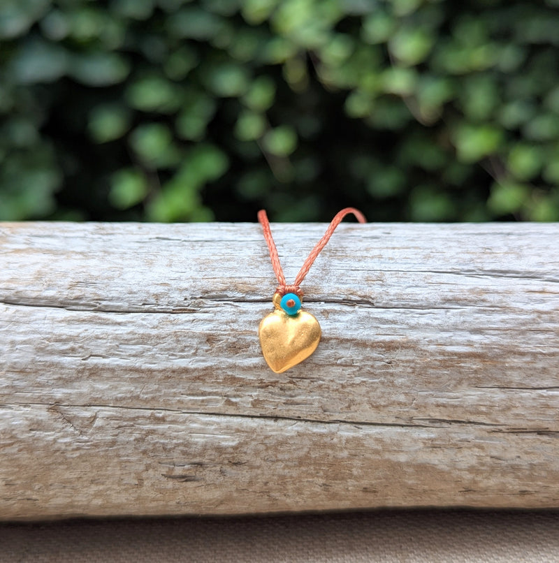 Gold heart-shaped pendant with a blue stone on an orange cord, resting on a wooden surface with a blurred green background.