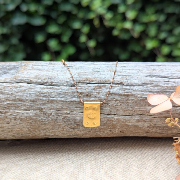 Gold necklace with a rectangular pendant with a textured design and a tan cord on a wooden log with a blurred green background