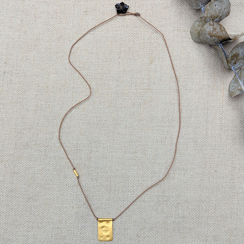 Gold necklace with a rectangular pendant on a tan cord with a black flower bead clasp on a textured beige surface