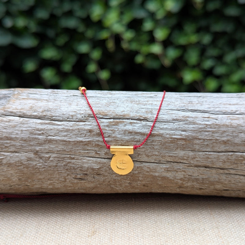 Gold circular pendent necklace with a red cord on a wooden log with a blurred green background