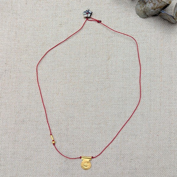 Necklace with red cord and gold circular pendant with a textured design and a black flower bead clasp on a textured beige surface