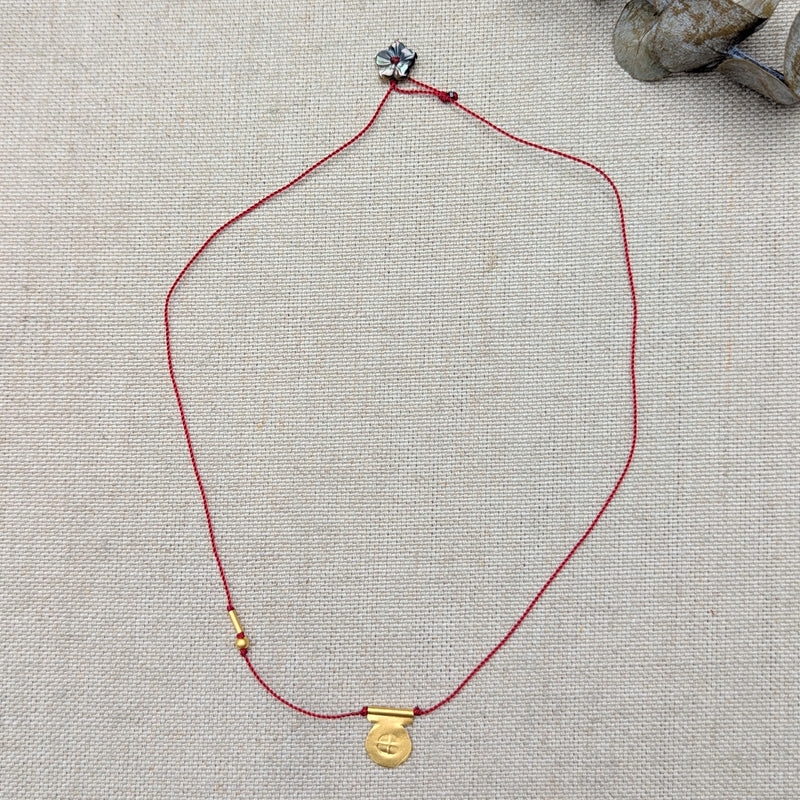 Necklace with red cord and gold circular pendant with a textured design and a black flower bead clasp on a textured beige surface